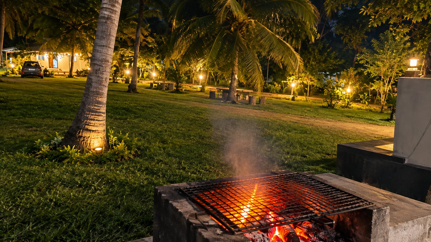 A charcoal barbecue glowing on the lawn at night, the garden lamps lit behind it